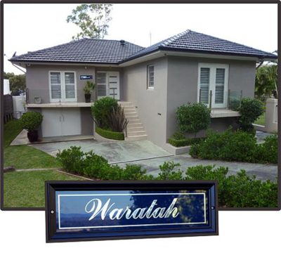 Custom black framed house sign reading 'Waratah' with elegant white script text, displayed over a modern grey two-story suburban home with a landscaped front yard