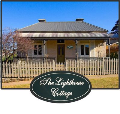 Classic oval timber house sign reading 'The Lighthouse Cottage' featuring a white cursive font on a dark green background, displayed in front of a historic heritage stone home.